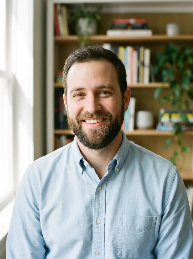 Natural headshot in a light-blue oxford shirt, softly blurred bookshelf background, natural window light, friendly expression